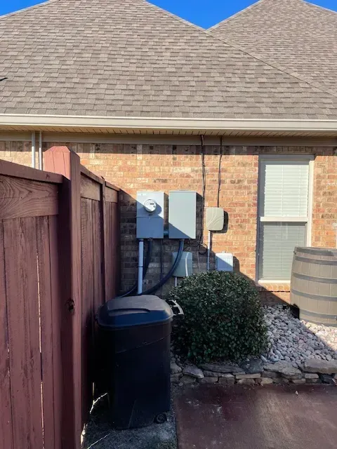 Electrical boxes on brick wall next to window, behind a red fence and black trash can.