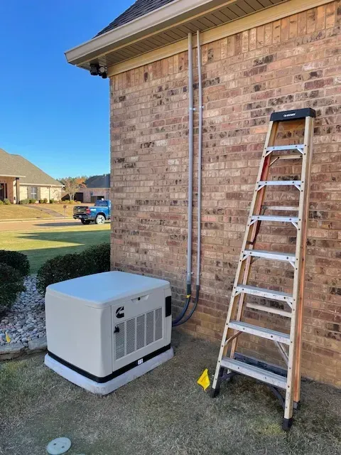 Generator next to a brick house. Ladder propped against wall. Conduit and blue sky.