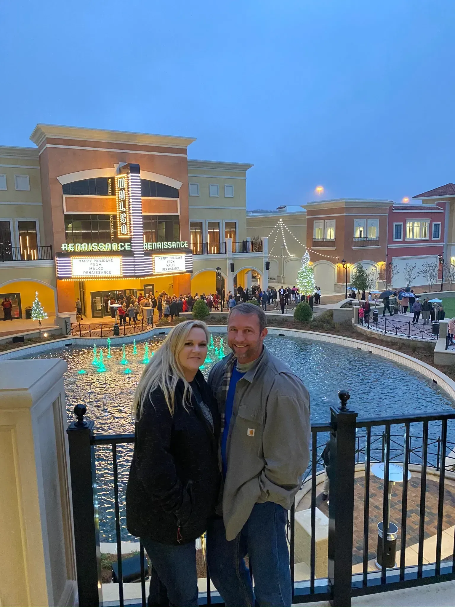 Couple posing by a fountain in front of a shopping center with lights and a Christmas tree.