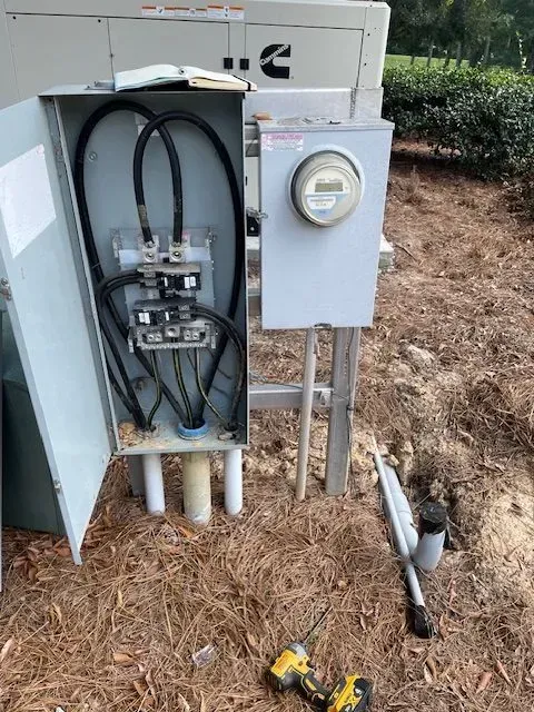 Electrical panel with open door, meter, and conduit, outside. Tools and ground visible.