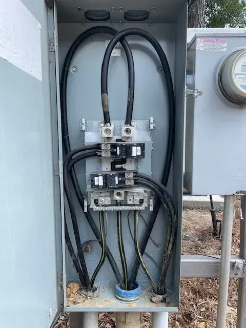 Open electrical panel with black wires connected to busbars, outdoor setting.