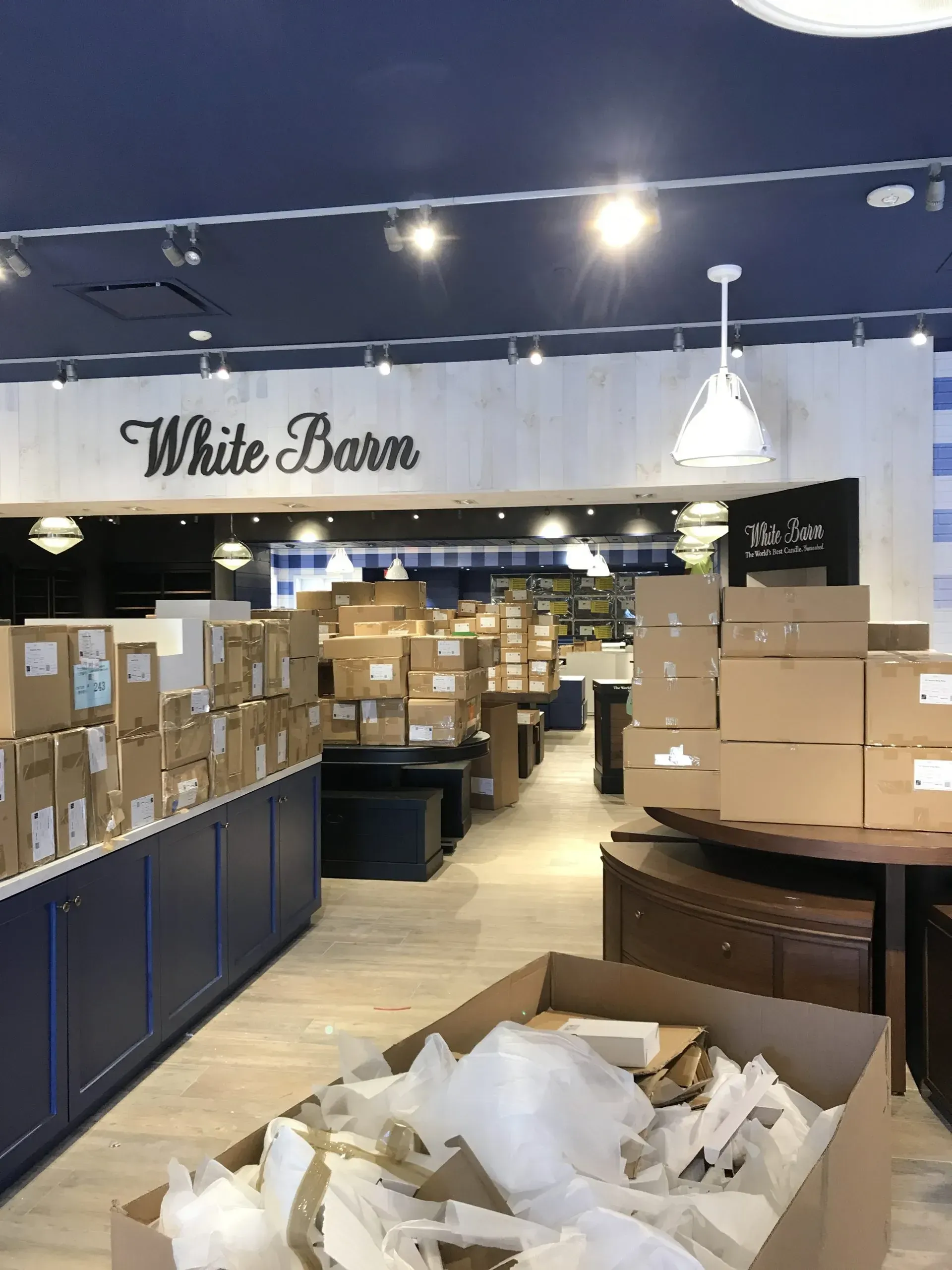 Inside a White Barn store, many cardboard boxes are stacked on display counters and the floor during a store reset.