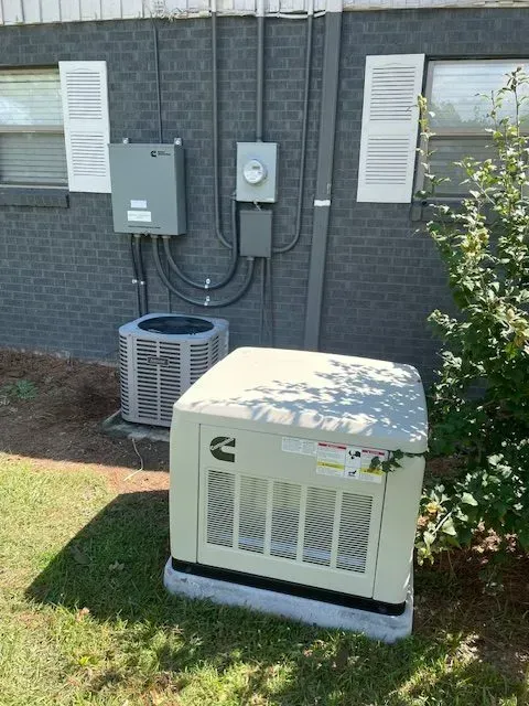 Outdoor electrical equipment, including an air conditioner and generator, next to a brick building.