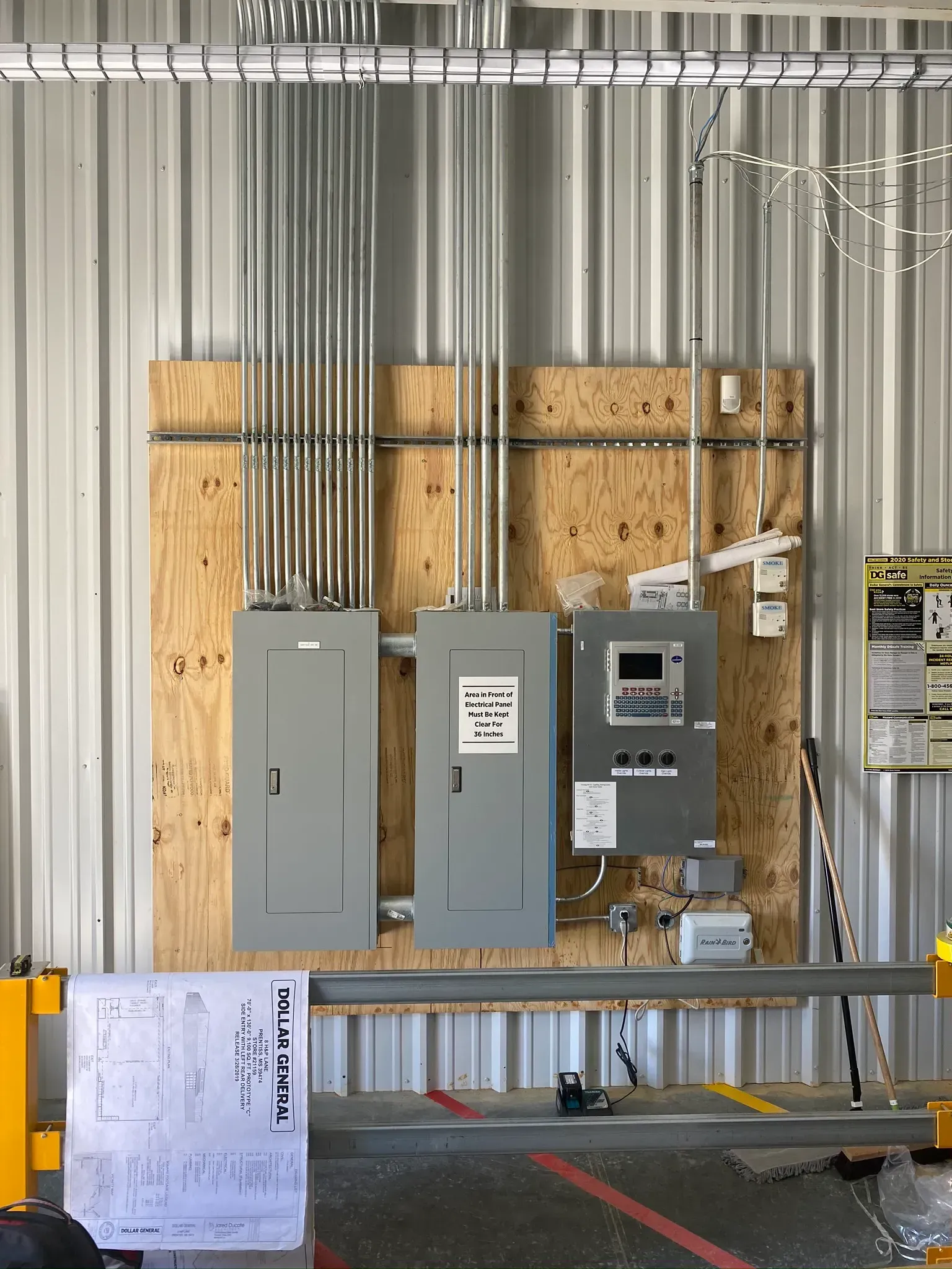 Electrical panels and conduits on plywood in a metal-walled industrial setting.