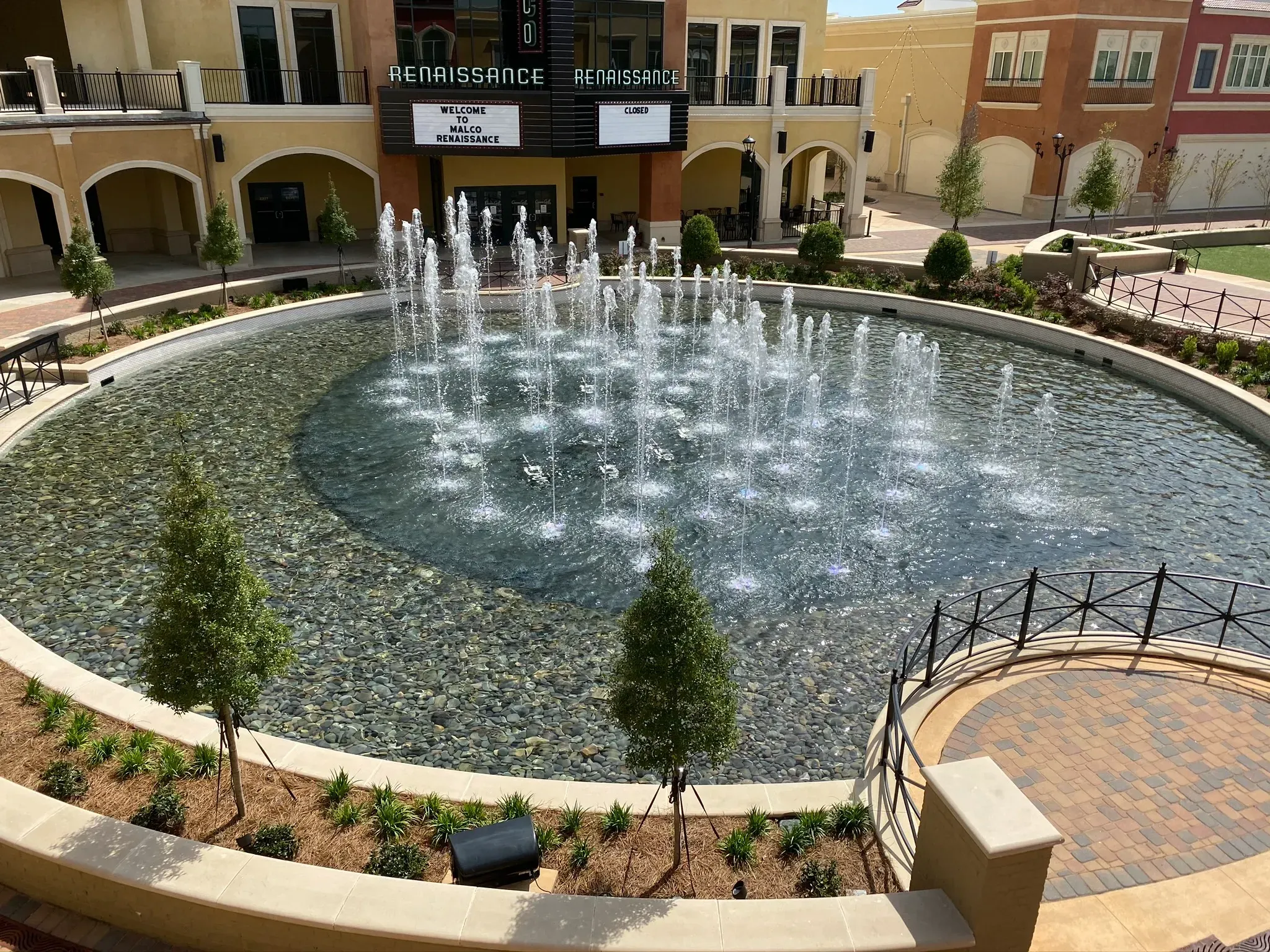 Circular fountain in a courtyard, spraying water; surrounded by trees, buildings, and a brick patio.