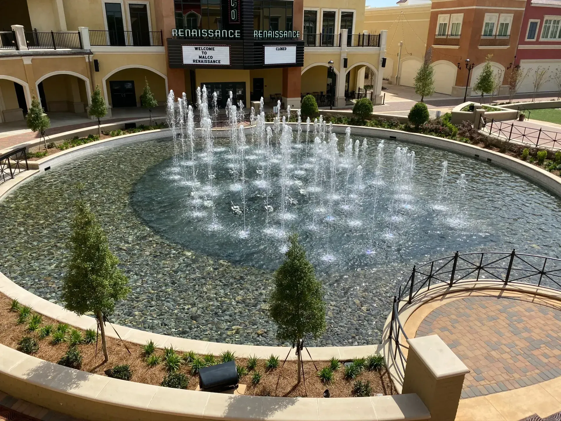 Fountain in circular pool at outdoor shopping center, water sprays upwards. Beige and tan buildings surround it.