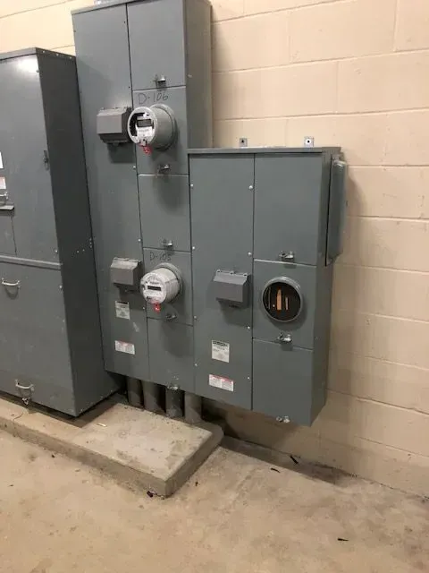 Electrical panel equipment on a concrete platform against a light-colored wall; gray metal boxes, meters visible.