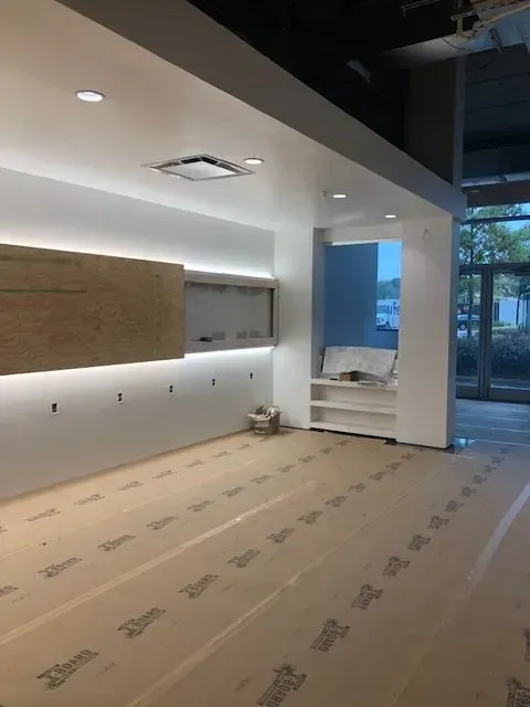 Interior view of a room under construction with white walls, recessed lighting, and protective floor covering.